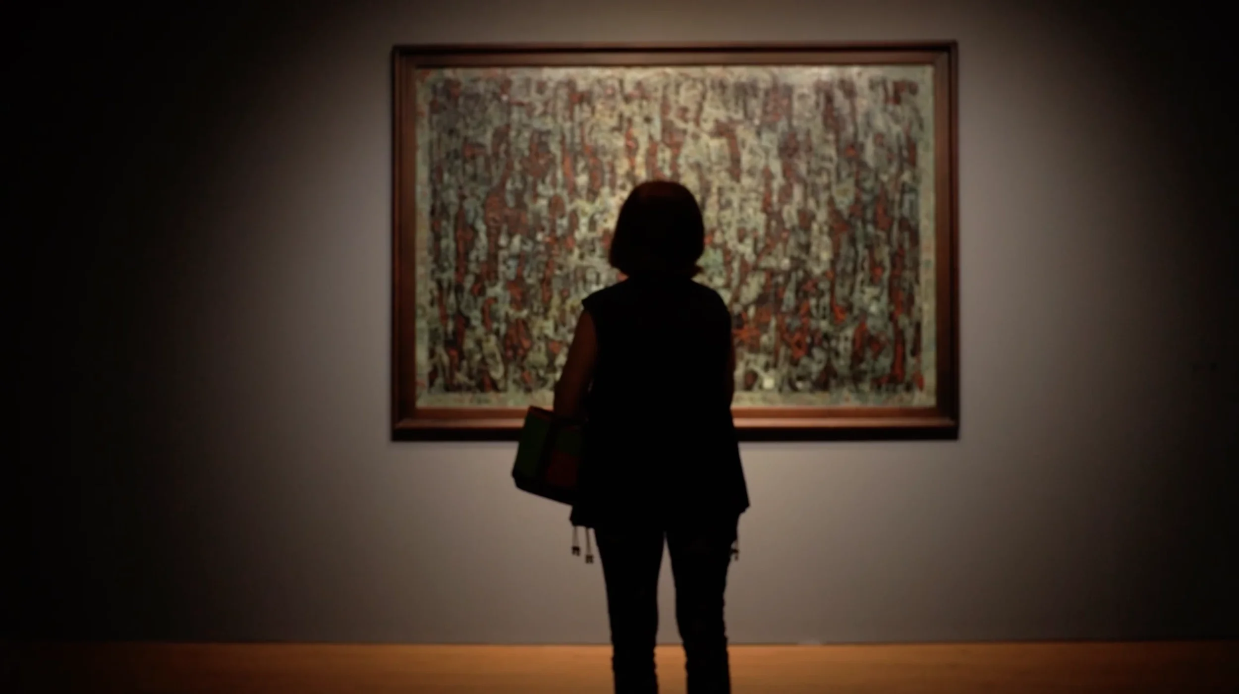 A person with dark hair and a black outfit looking at a framed artwork in a dimly lit art gallery.