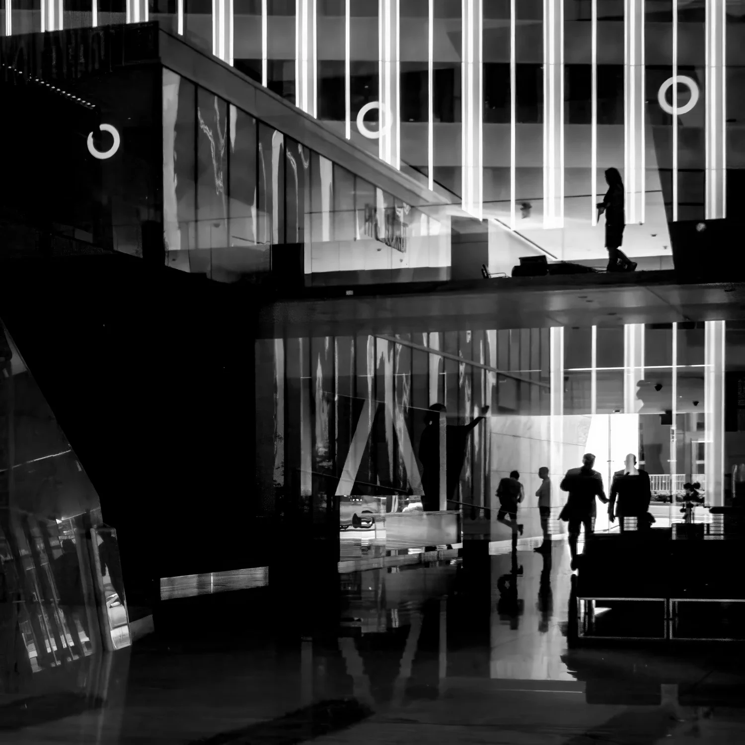 Silhouettes of people inside a modern, illuminated building with glass walls and vertical light fixtures, reflected on a shiny floor.