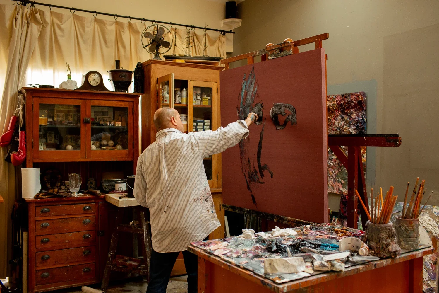 An artist working on a large canvas in his studio, which is filled with art supplies, wooden cabinets, and paintings.