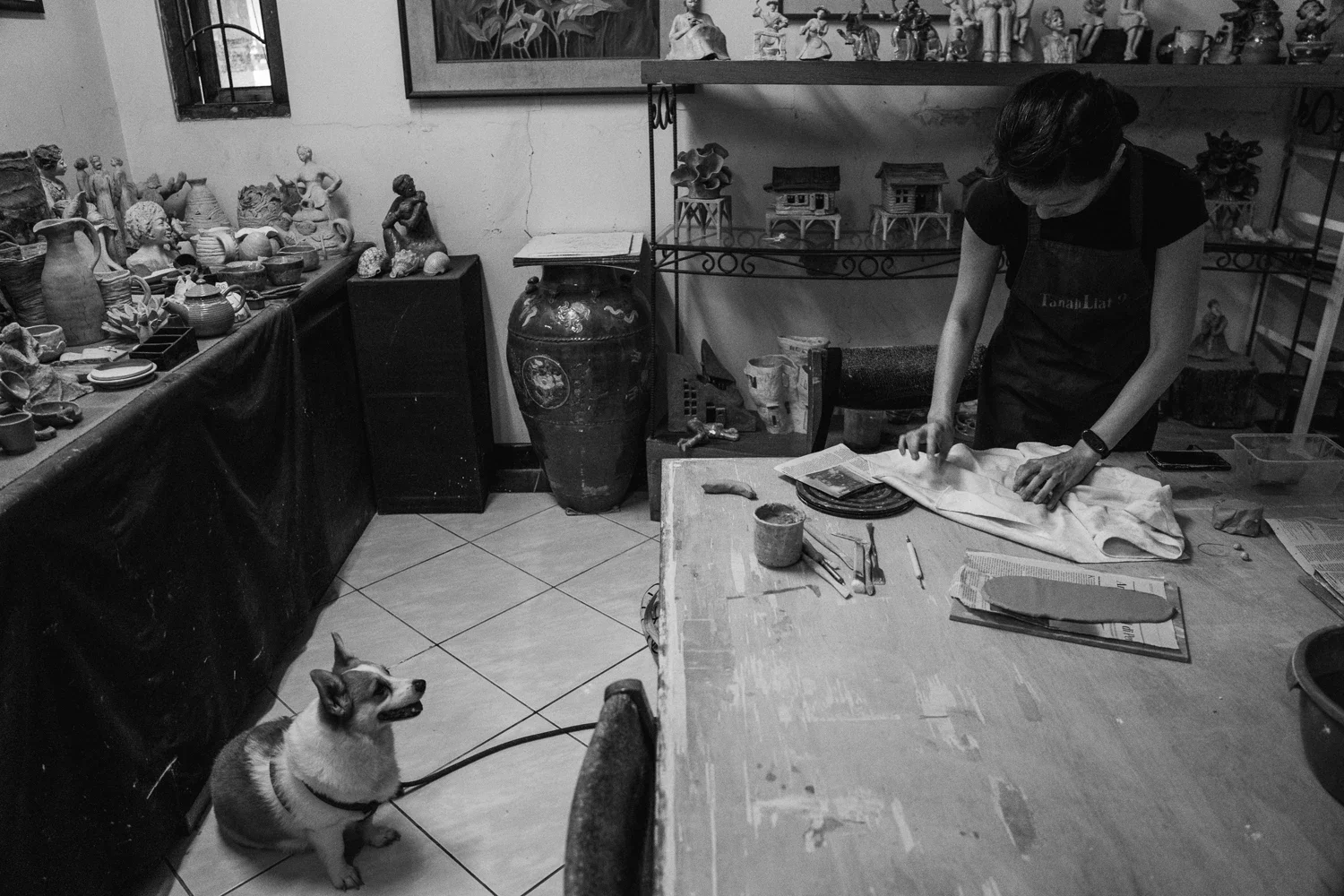 A woman working on fabric at a wooden table in an art studio, with a dog sitting on the tiled floor nearby, surrounded by shelves and tables with various sculptures and pottery.