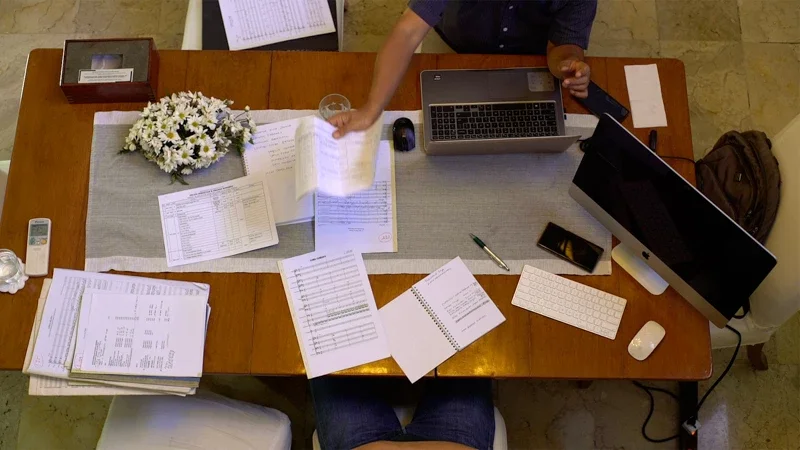 Overhead view of a cluttered wooden desk with papers, a laptop, a monitor, a coffee glass, a flower arrangement, a pen, a keyboard, a mouse, a remote, and a backpack, with a person working at the desk.