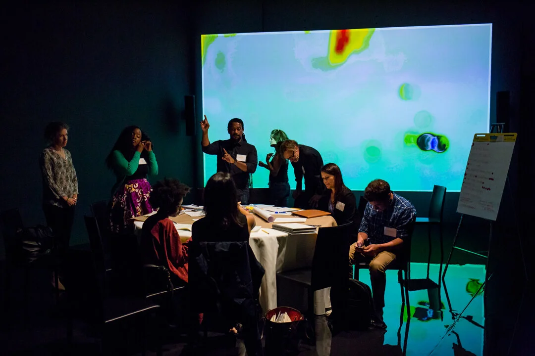 9 people working and discussing an important matter around a table filled with notes and papers, in a dark room with a colorful arty projection as their background