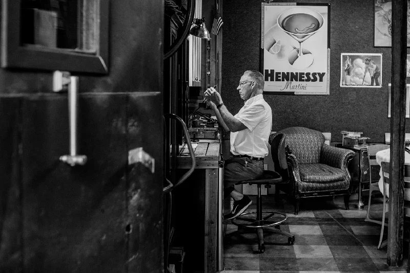 Kenton Nelson sitting at his work bench, working on a project in a room with vintage decor and framed artwork on the wall.