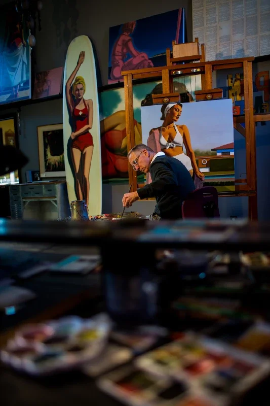 An artist in a dark art studio or gallery, working on a painting of a woman with a paddle board and blue sky, surrounded by completed artworks, with art supplies in the foreground.
