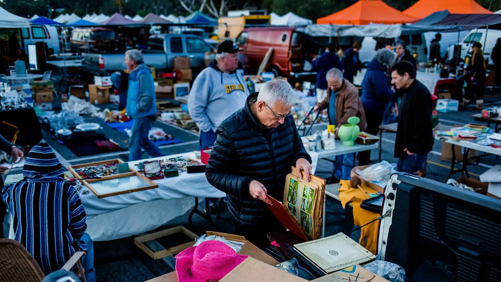 Jim Heimann browsing an old photo album at the Pasadena Swap Meet, surrounded by people looking and walking around amongst tables of items.
