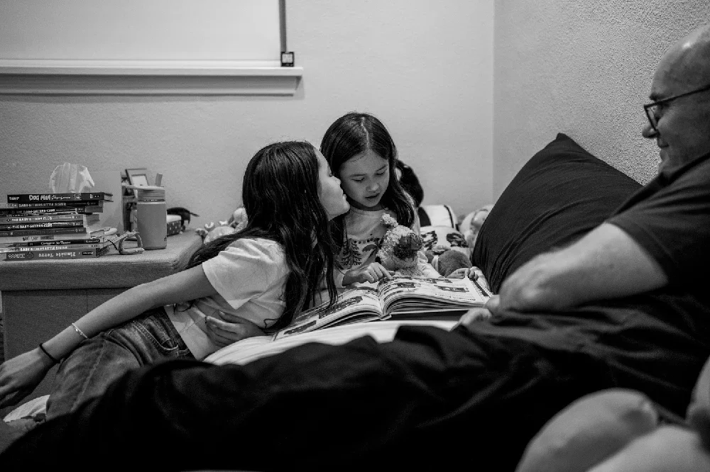 Two young girls and an adult man are sitting on a bed, looking at a book. The girl in the middle is holding a small stuffed animal, and the girl on the left is kissing her on the cheek. The room has a cluttered desk with books and other items.