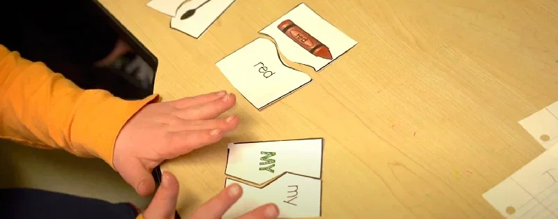 Child’s hands arranging word puzzle pieces with words 'red' and 'my' on a wooden table.