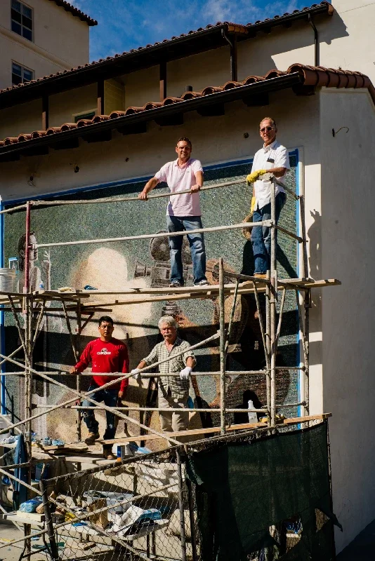 Kenton Nelson and his crew on scaffolding working on the pasadena playhouse mosaic mural on the side of a building under a blue sky.
