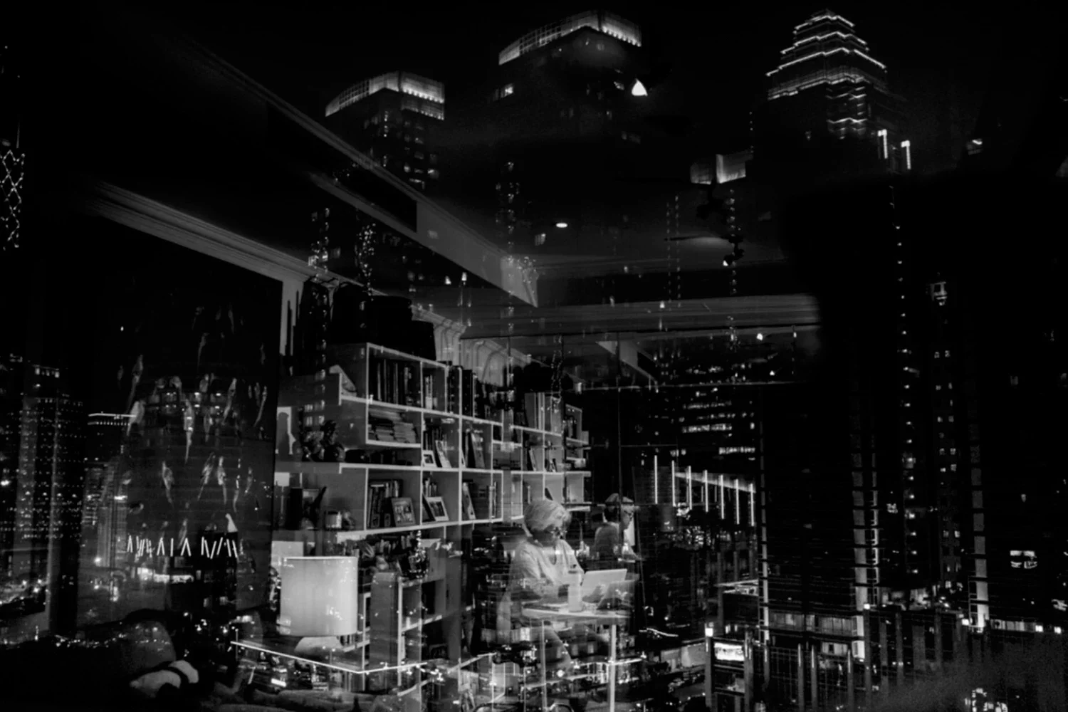 Nighttime cityscape reflected in a window showing an interior with bookshelves, a woman working at a table, and illuminated high-rise buildings outside.