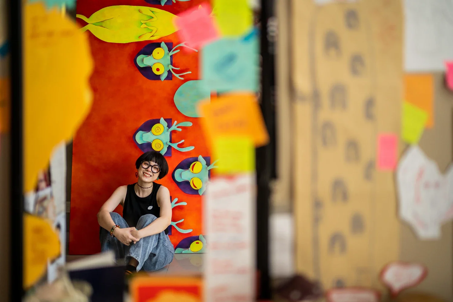 A person with short dark hair, glasses, and a black sleeveless top, smiling and sitting cross-legged in front of a colorful backdrop with animal characters
