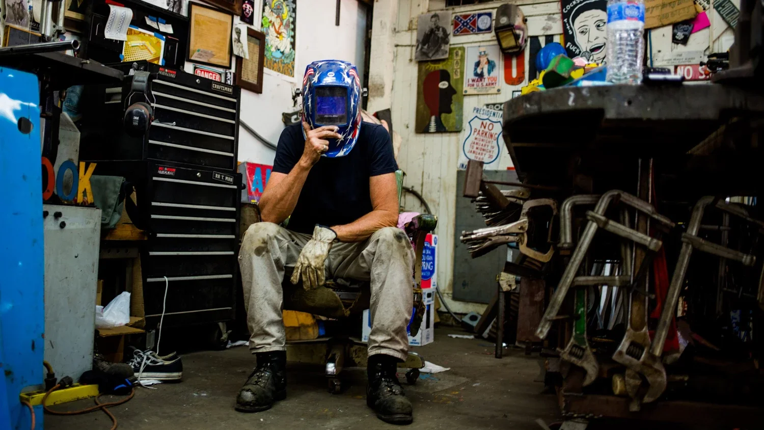 Metal Sculptor David Buckingham sitting on a stool in a cluttered workshop, wearing a protective welding helmet, black shirt, and beige pants, surrounded by tools and various signs on the walls.