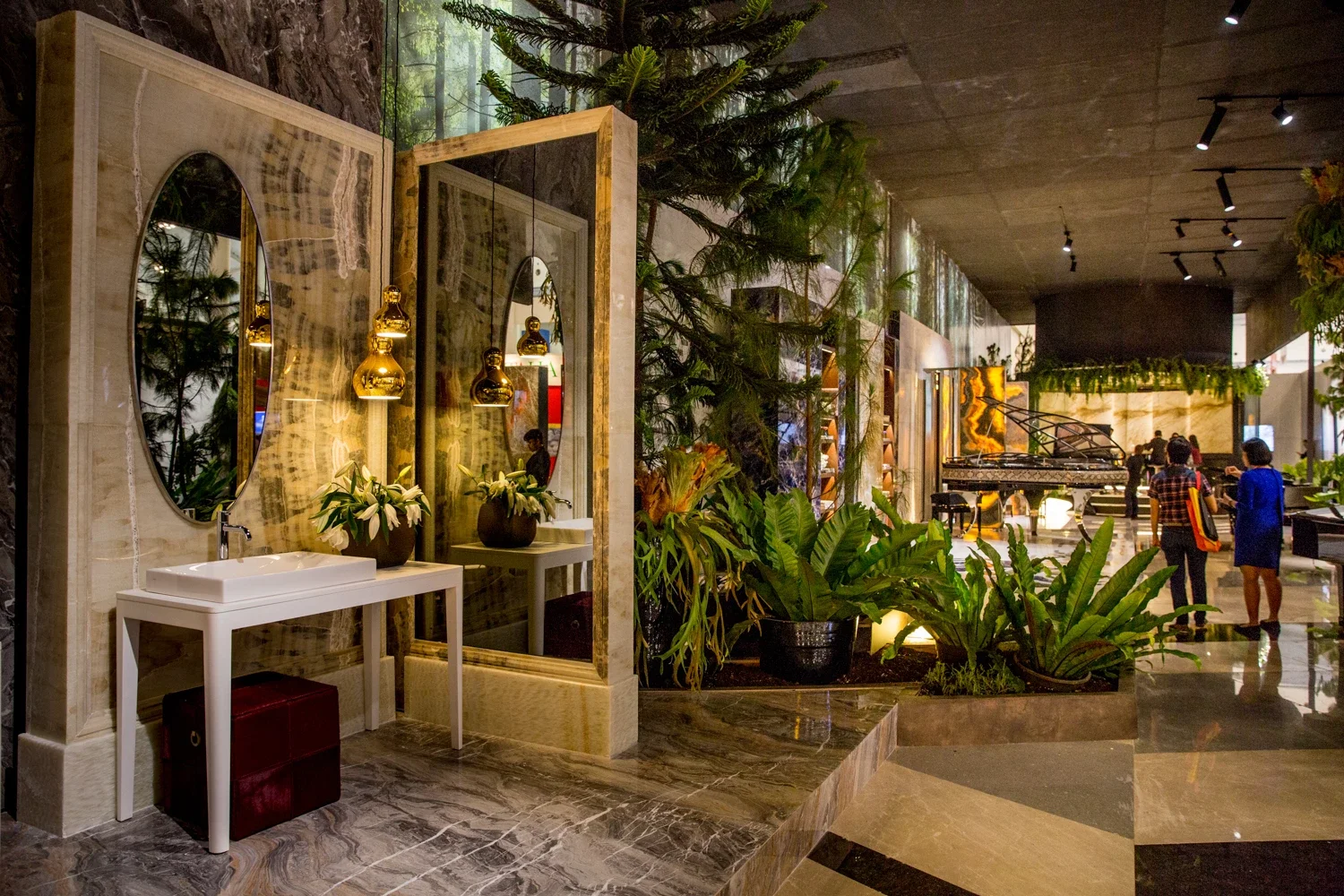 Interior of a hotel lobby with large mirrors, white sinks, gold pendant lights, lush green plants, and a grand piano in the background. People are walking and socializing.