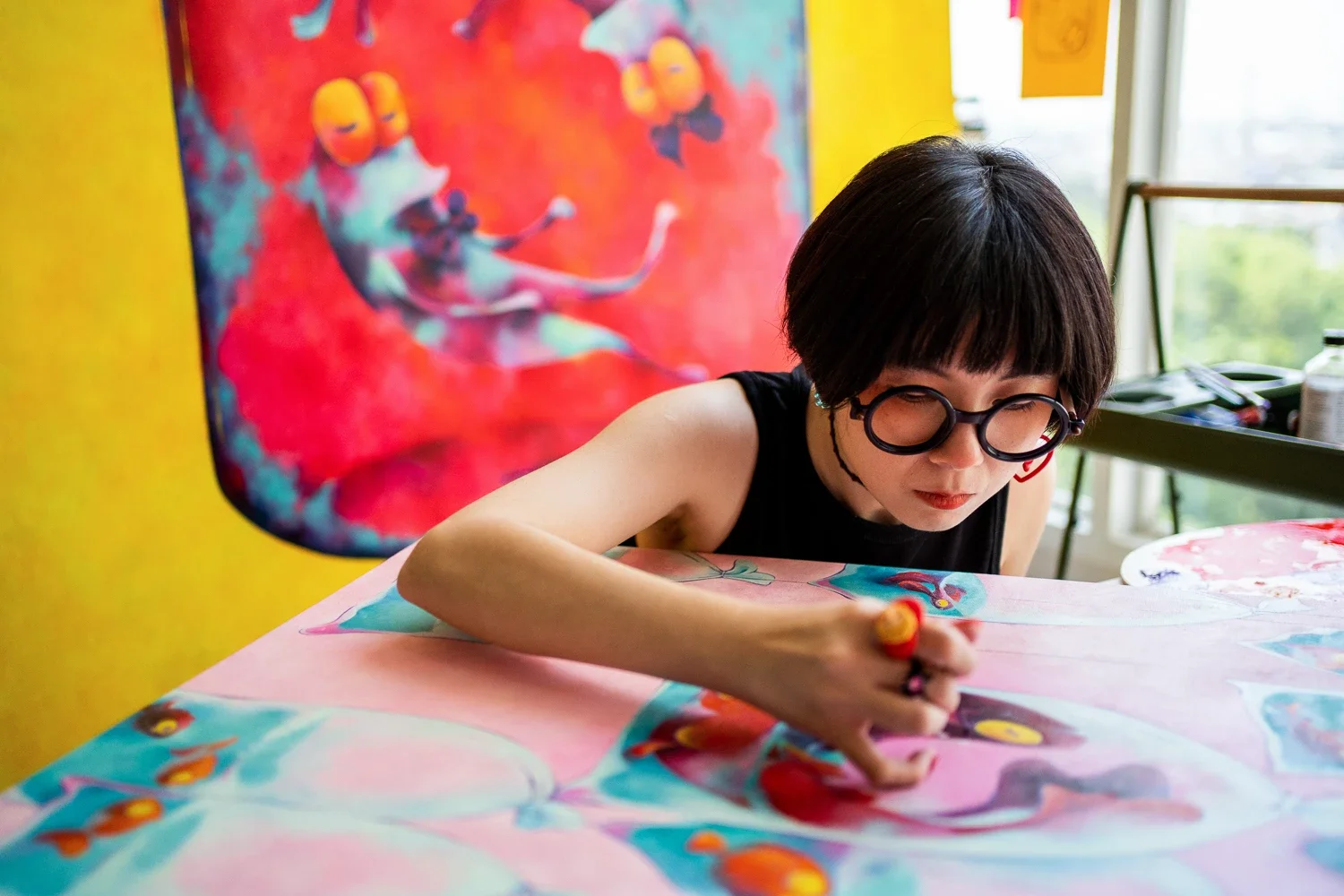 A woman with dark hair, glasses, and a black sleeveless shirt is painting abstract art with bright colors on a large canvas. Behind her is a colorful painting on the wall and a window letting in natural light.