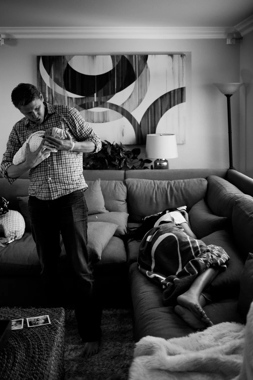 A man holding a newborn baby in a living room with a woman sleeping on the couch, a lamp, a large abstract painting on the wall, and some photos on a table.