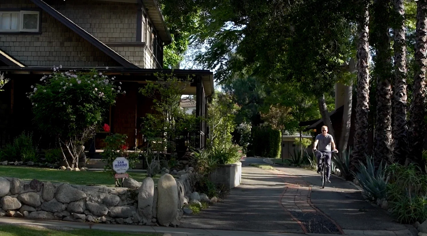A man riding a bicycle on a sidewalk lined with tall palm trees and lush greenery in front of a house with a stone wall and garden.