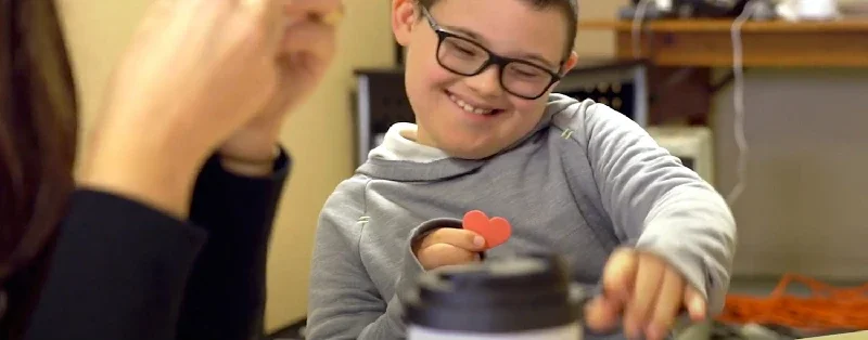 A young boy smiling and holding a small heart-shaped object, sitting at a table with a woman who is partially visible.