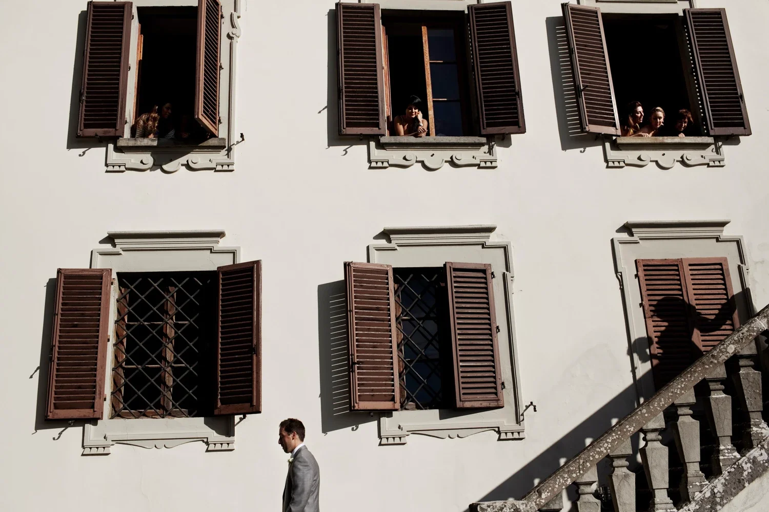 A building with six windows with open shutters, three on the top row and three on the bottom row. Four women are leaning out of the top right window, two at the left window, and a man in a gray suit is walking past the building. Shadows of the open shutters are cast on the white wall.