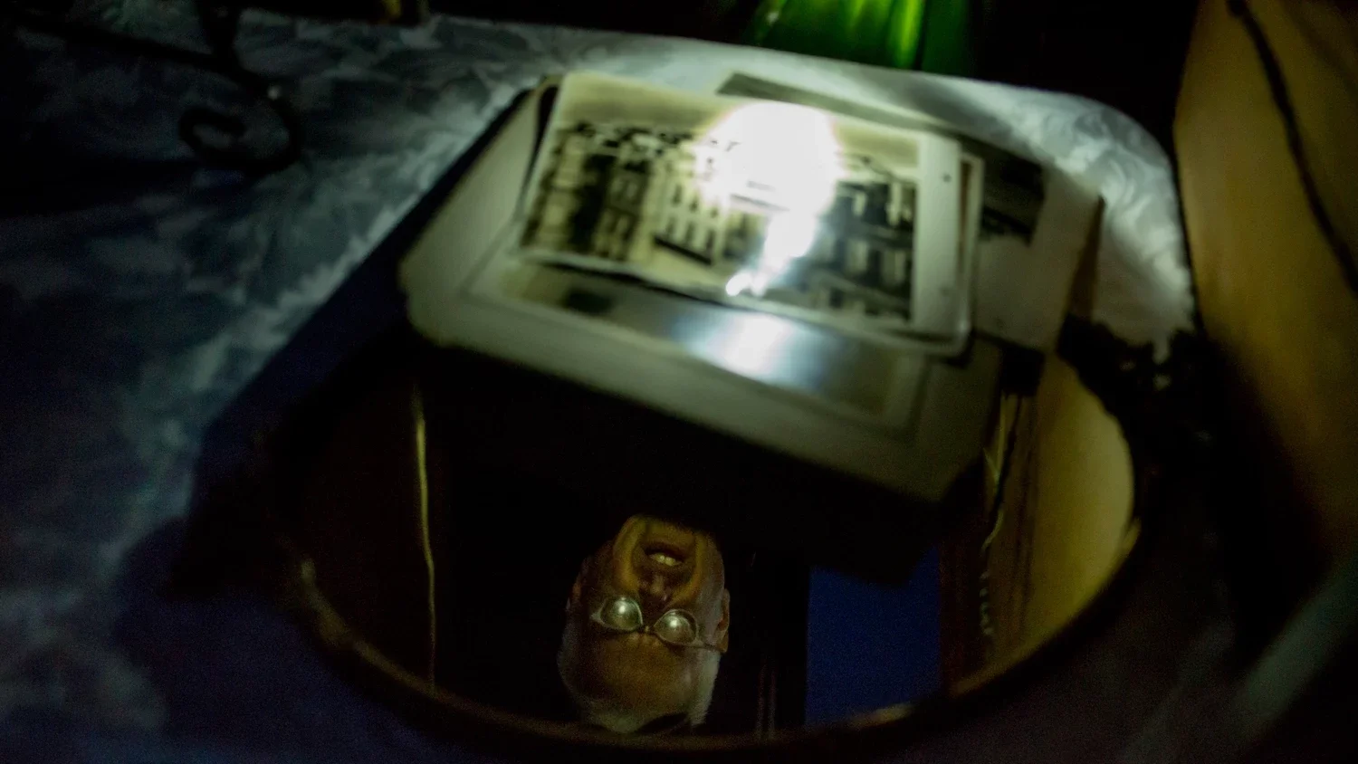 Reflection of a man wearing glasses and smiling, taking a photo with a phone, in a mirror reflecting a bookshelf and a lamp.