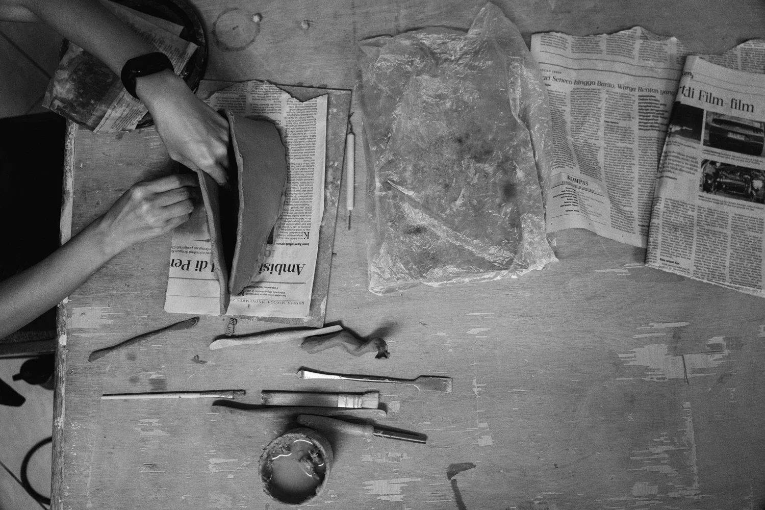 Person working with clay on a wooden table, surrounded by newspapers, sculpting tools, and a plastic bag with more clay.