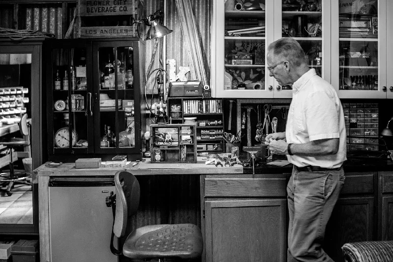 Artist Kenton Nelson working in a workshop or craft room filled with tools, storage cabinets, and various supplies.