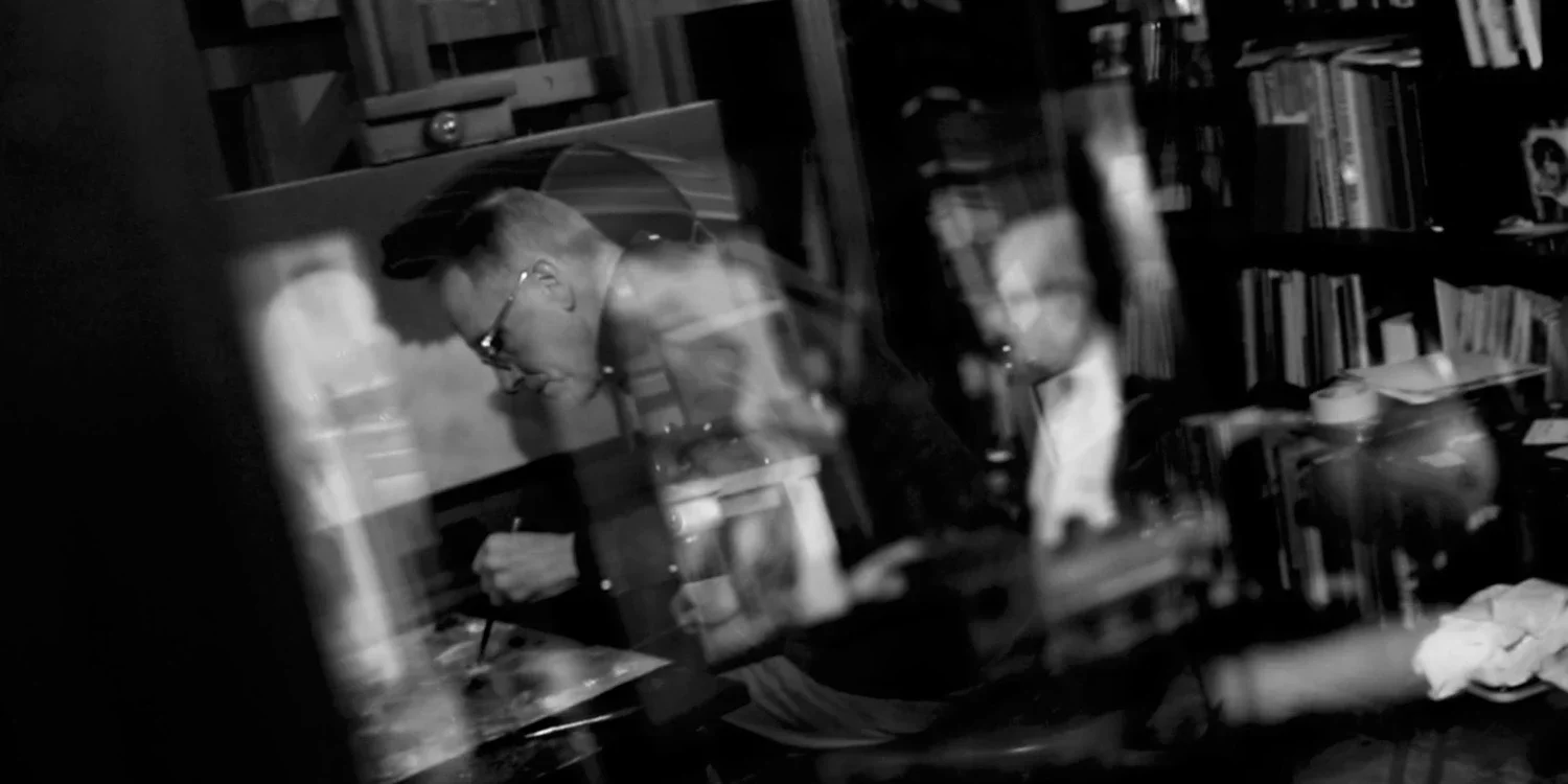 Black and white photo of a man in glasses sitting at a desk, writing or drawing, with a bookshelf filled with books in the background, and reflections on a glass surface in front.