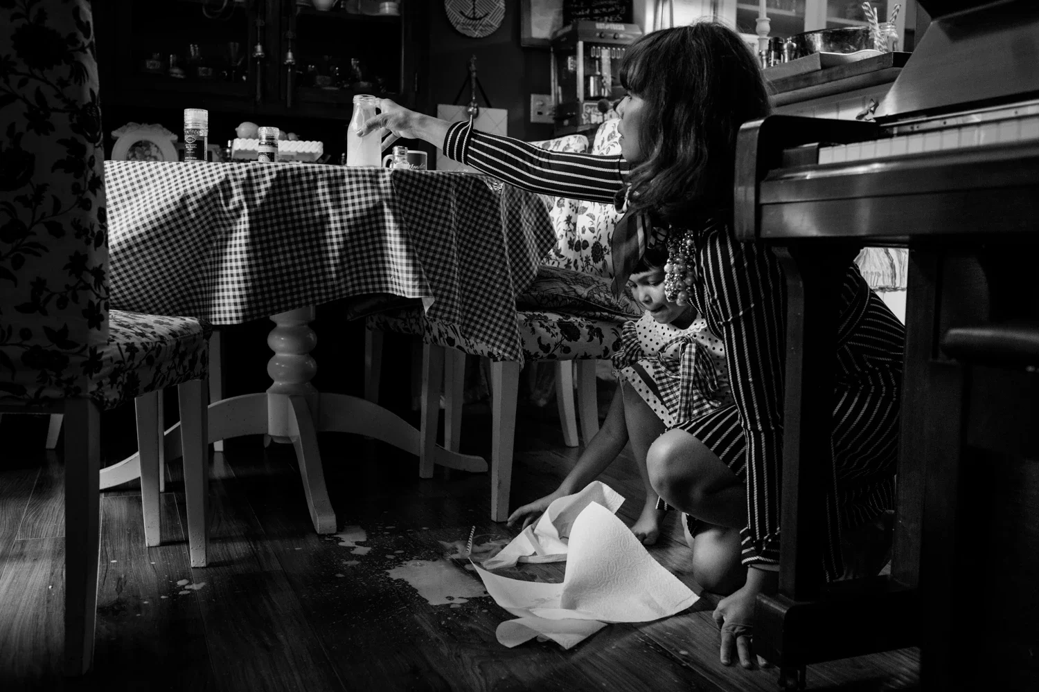 A woman and a young girl are underneath a dining table with a checkered tablecloth. The woman is reaching out, possibly trying to clean or pick something up, while the girl looks on. There is a spilled liquid and paper towels on the floor, indicating an accident or spill.