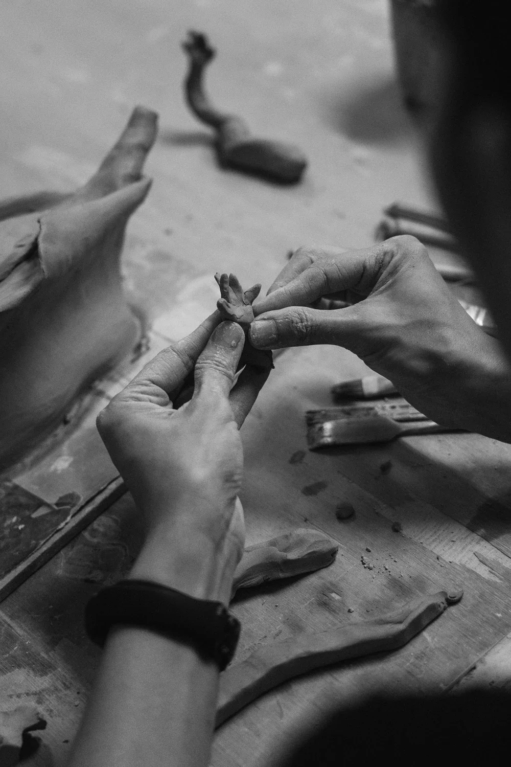 Person working on a clay sculpture, shaping a small figure with their hands, surrounded by clay tools on a wooden surface.