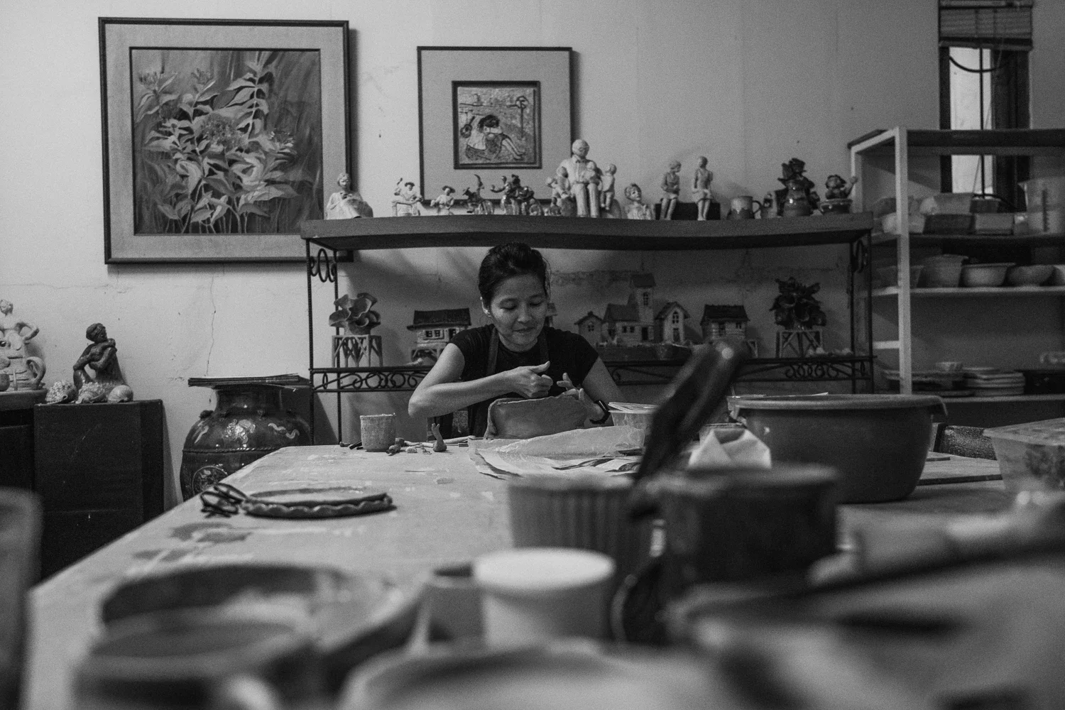 A woman is sitting at a table in a room decorated with various framed artwork and figurines, looking into her bag with art supplies around her.