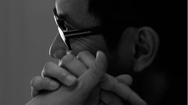 A person wearing glasses is holding their hands together near their face, with a contemplative expression.