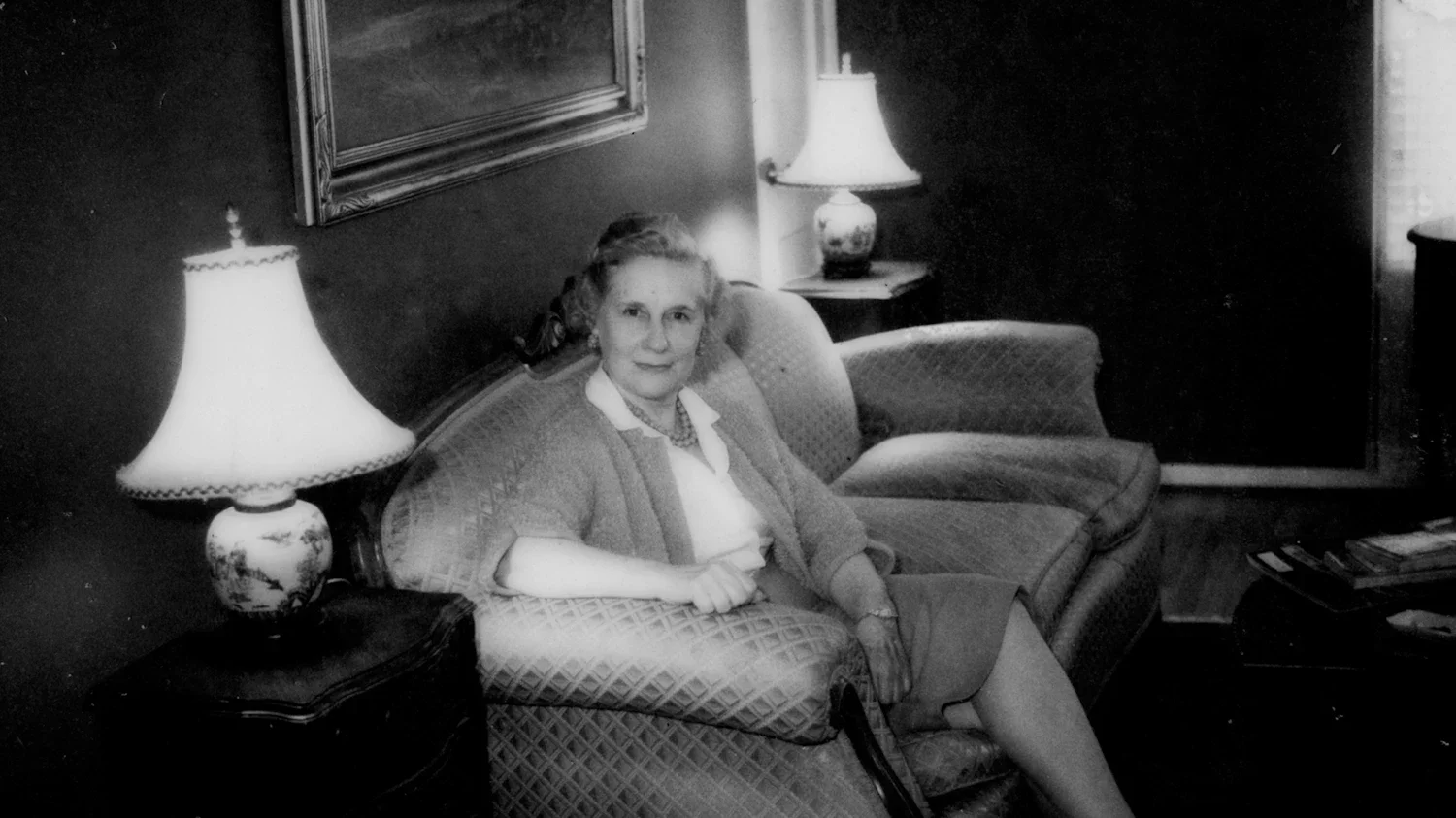 A black and white photo of a woman sitting on a patterned sofa in a living room, with a lamp on a side table next to her, and framed artwork on the wall behind.