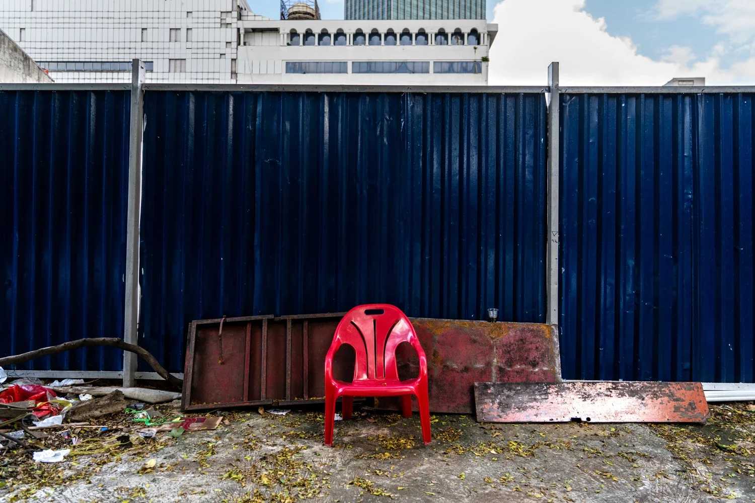 A red plastic chair placed on the ground in front of a blue corrugated metal fence, with scattered debris and fallen leaves on the ground.