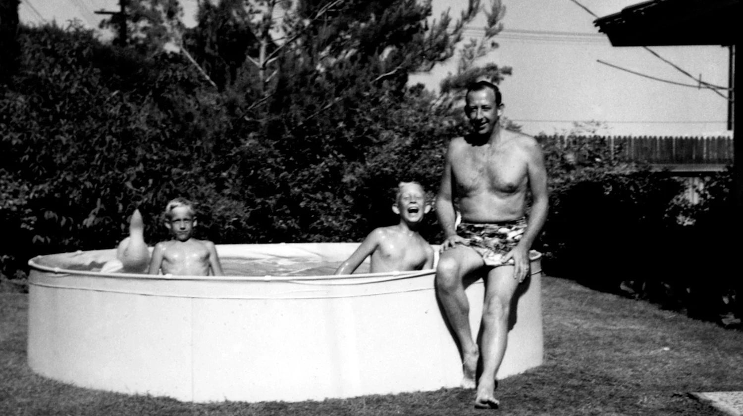 A black and white photo of three children and an adult in an outdoor hot tub, with trees and a fence in the background. The adult is sitting on the edge of the hot tub wearing swim trunks, and the children are in the water, smiling and playing.