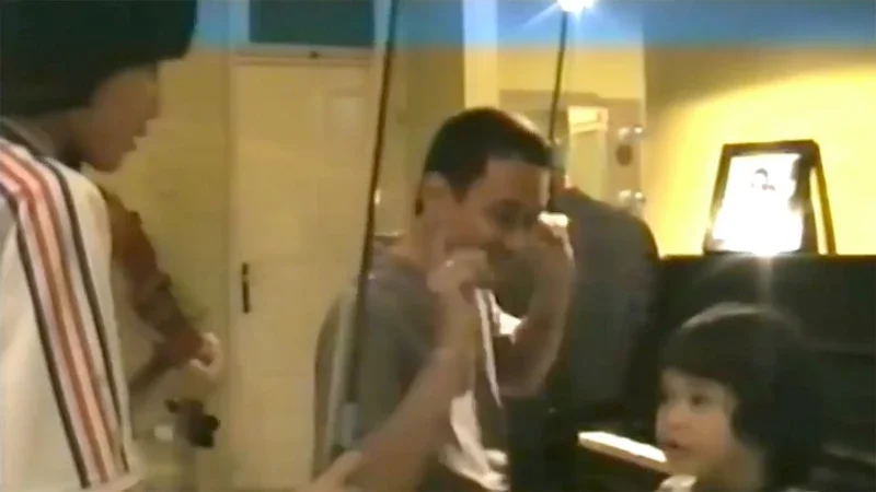 A woman standing next to a seated young boy with short dark hair, in a room with yellow walls and a bright light on a computer monitor in the background.