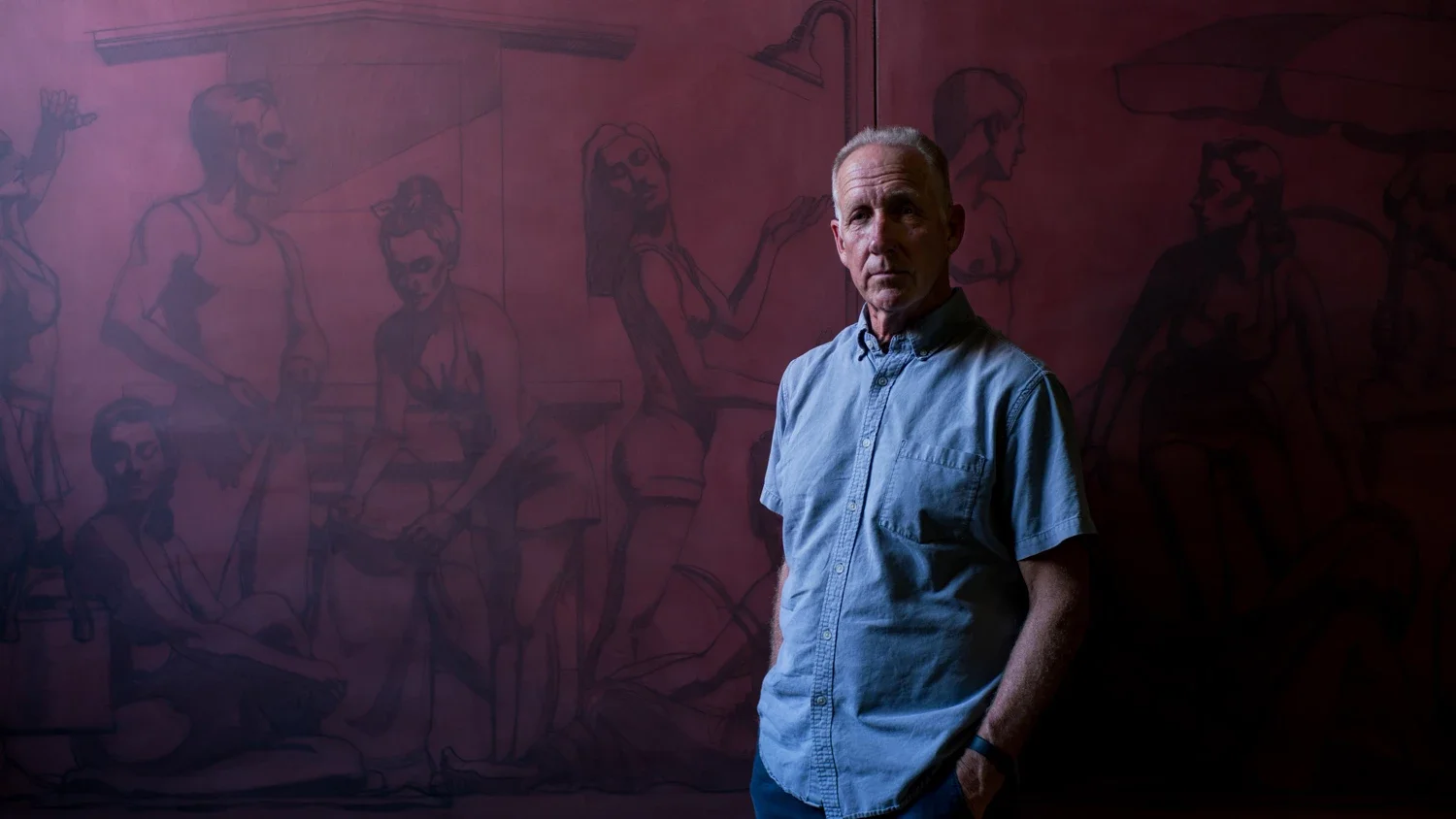 Artist Kenton Nelson in a light blue button-up shirt stands with his hands in his pockets in front of an unfinished painting depicting multiple outlined figures of people.