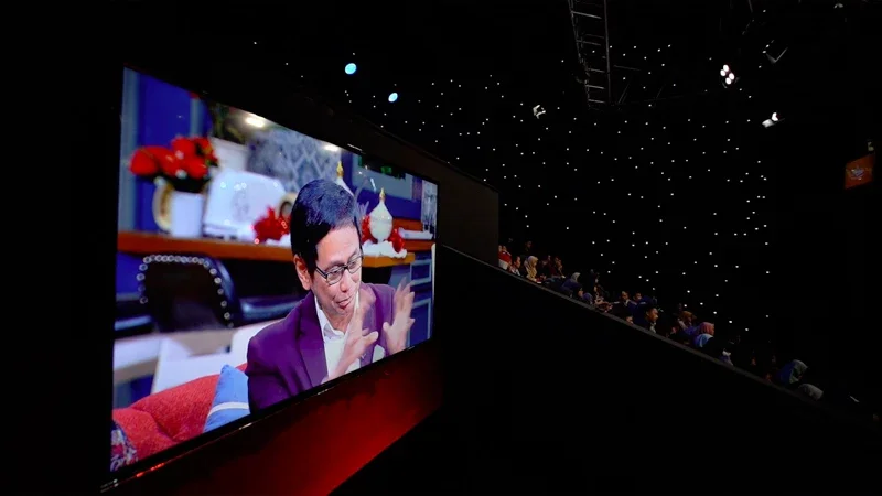 A large screen showing a man in glasses wearing a purple jacket, sitting in a decorated room. The room has Christmas decorations, including a small Christmas tree and red ornaments. The auditorium is filled with people watching the presentation, with a starry ceiling overhead.