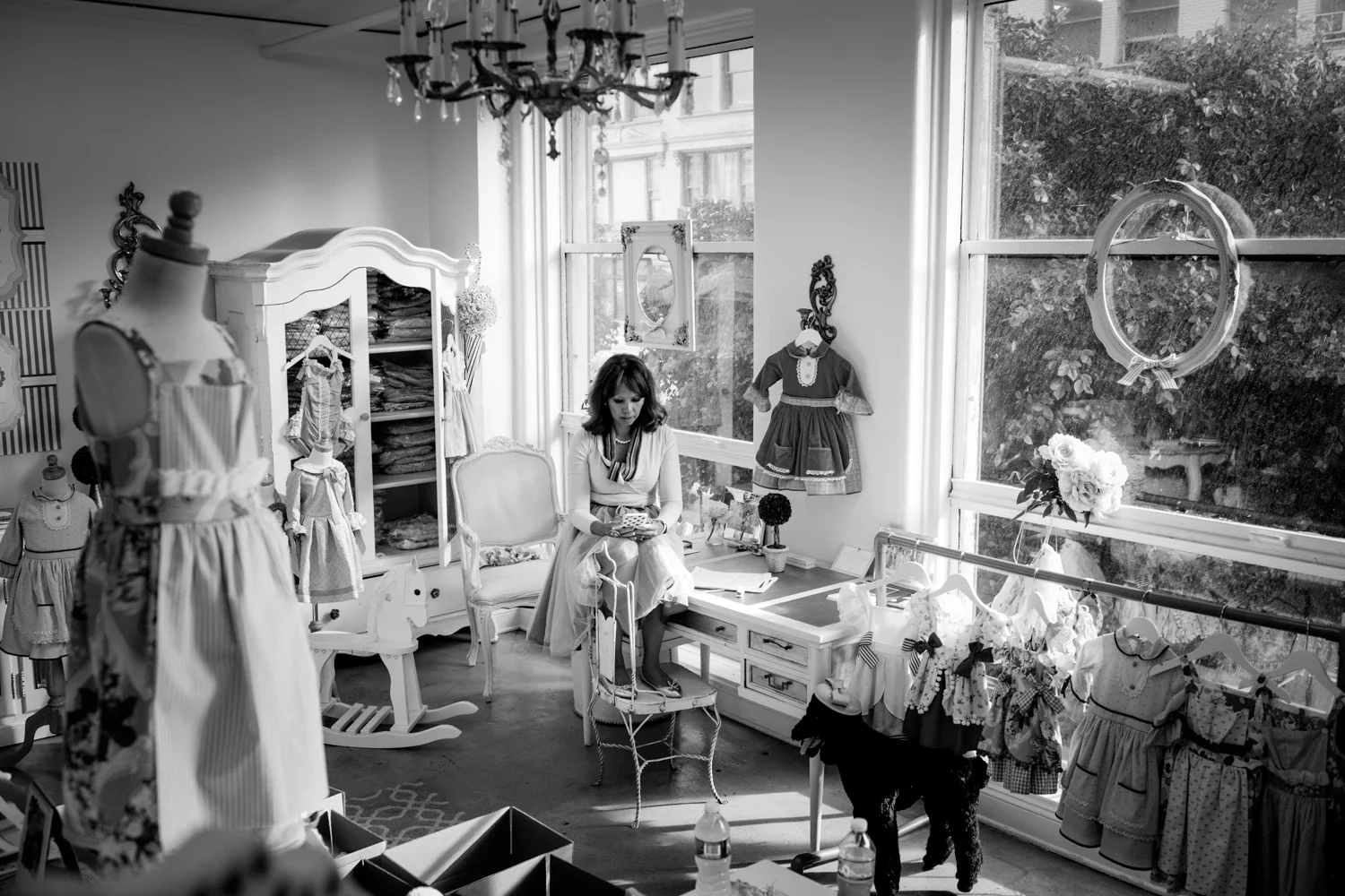 A woman sitting on a chair in a room filled with children's clothing and decorations, with large windows and a chandelier overhead.