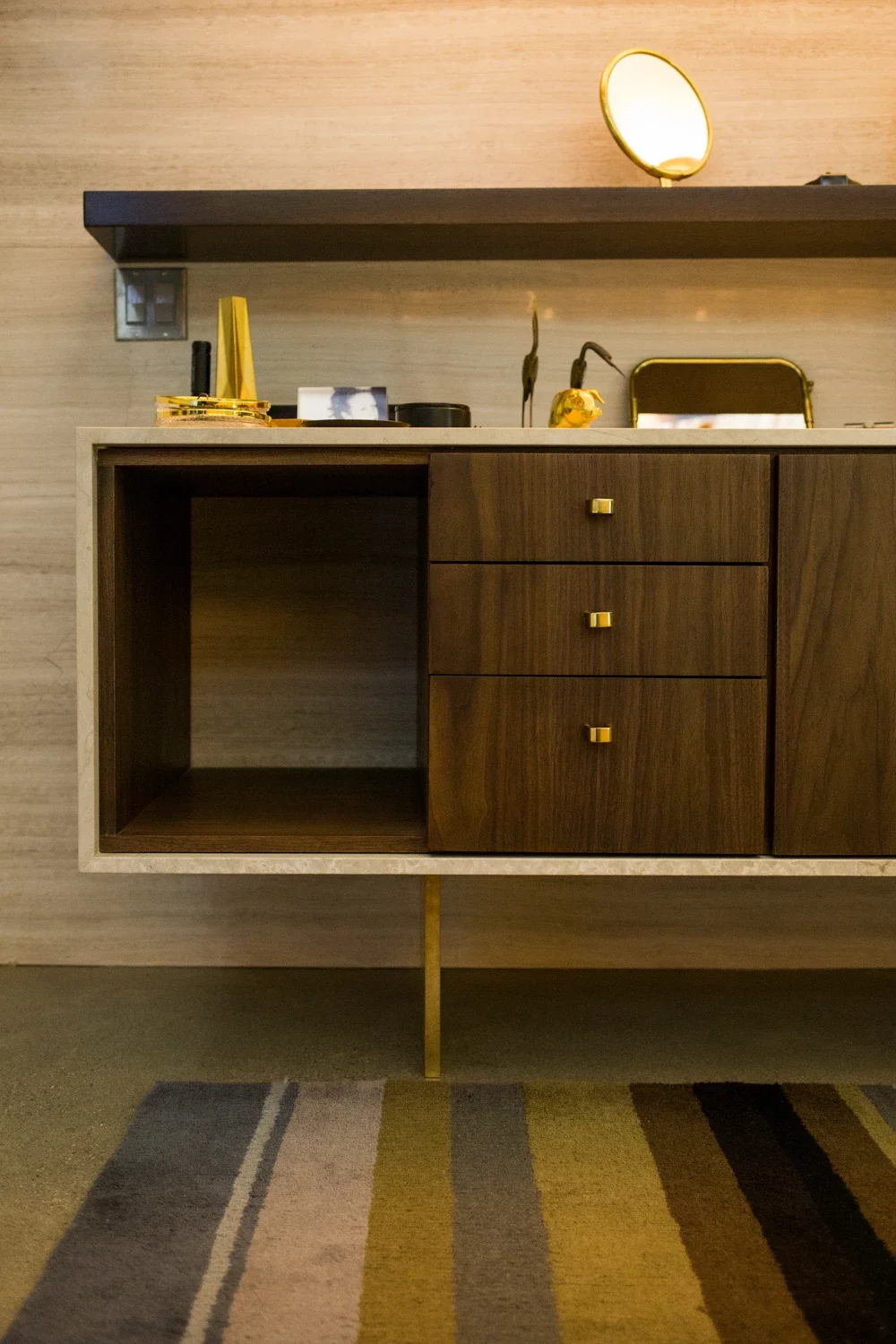 A mid-century modern sideboard with open and closed storage, placed against a beige wooden wall, with decorative items on top, including a small sculpture, a tray with yellow objects, and a mirror above.