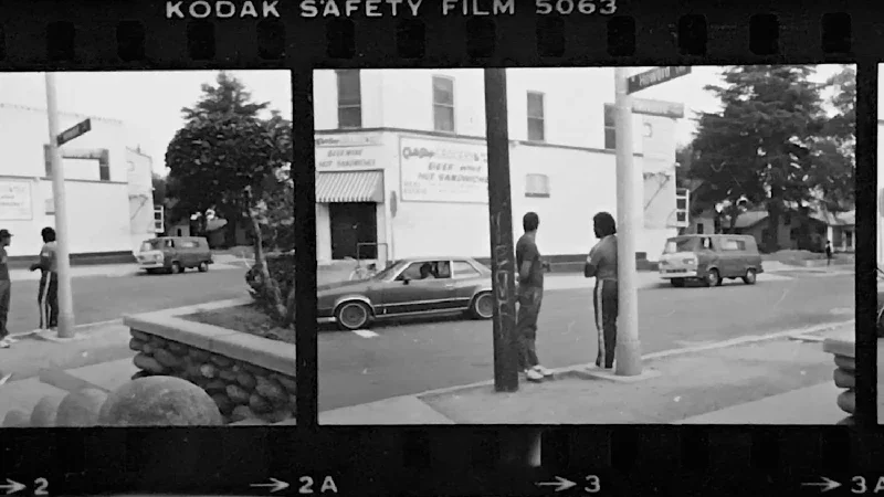 Black and white photo of a city street scene with two people conversing on the sidewalk, cars driving by, and buildings in the background.