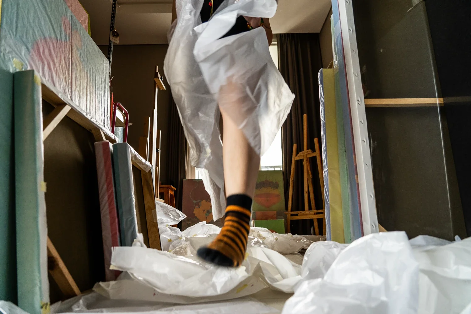 A person standing on a cluttered floor surrounded by crumpled paper, with books, painting supplies, and art materials in a room with large windows and wooden furniture.