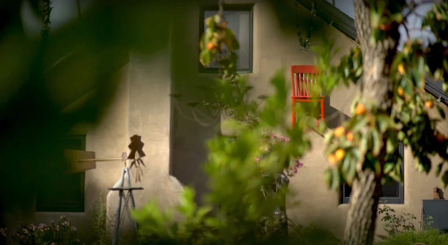 A red chair on an hanging high on the side of a building, partially obscured by green foliage and trees, with a window and wall in the background.