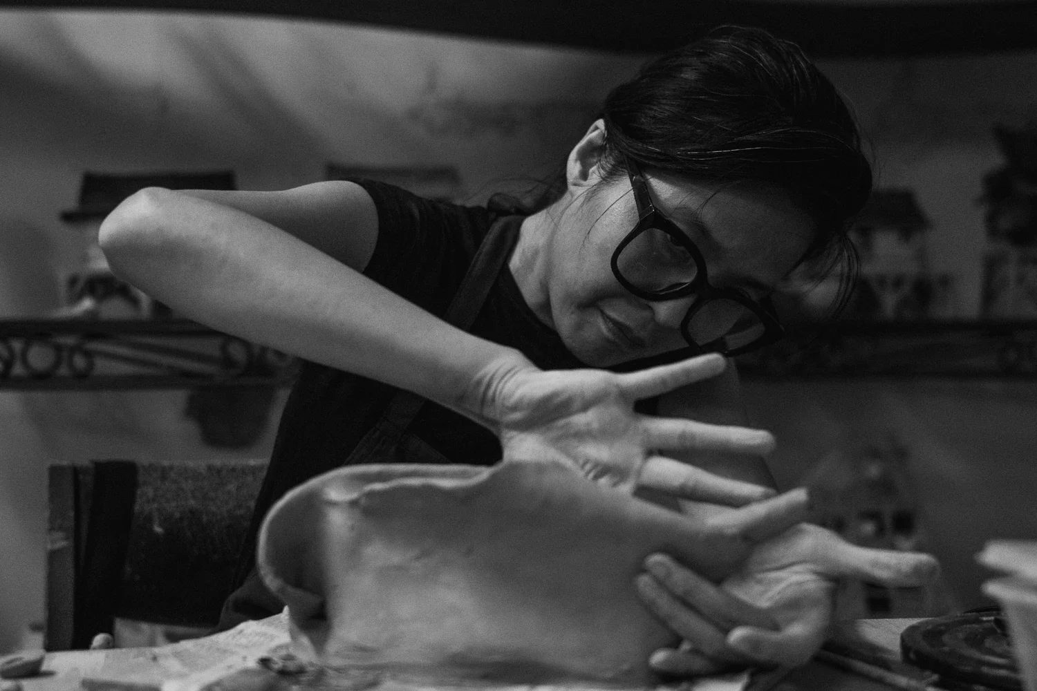 A woman with glasses is shaping or sculpting a large object, possibly clay or dough, on a table in a workshop or studio. She has dark hair and is focused on her work.