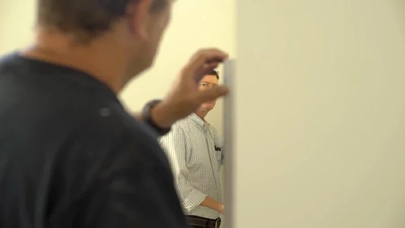 A person in the foreground gesturing towards a man standing behind a wall in an indoor setting.