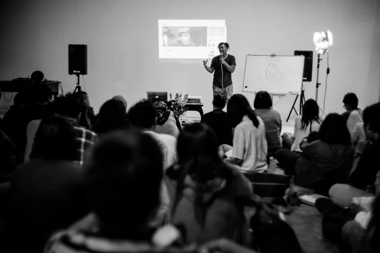 A speaker standing in front of an audience, giving a presentation. There is a projection on the wall showing a person's face, a whiteboard with a drawing, and audio speakers on either side.
