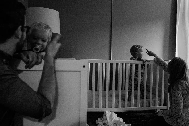 A man takes a photograph of a young child in a crib, while another child and woman interact outside the crib in a room.