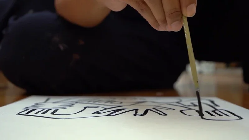 A person sitting on the floor, painting a large, black, abstract design on a white sheet of paper or canvas with a thin brush.