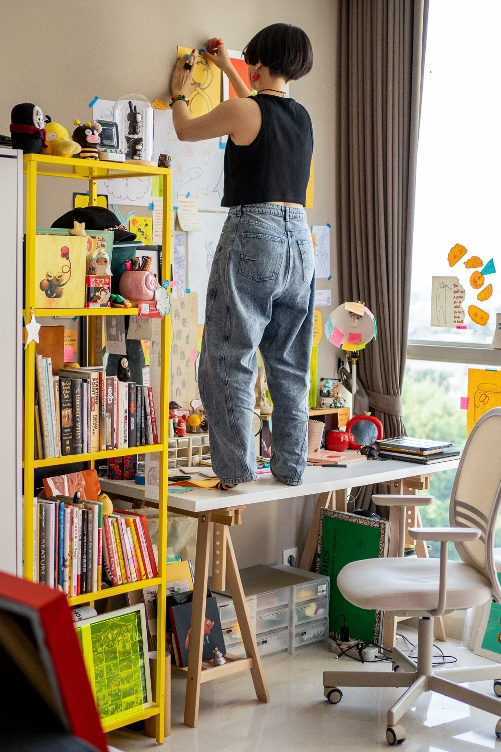 A woman with short dark hair wearing a black sleeveless top and gray jeans stands on a white desk, drawing on a wall with colorful art supplies. The room has a yellow bookshelf filled with books and toys, and various artworks and notes are taped on the wall and window.