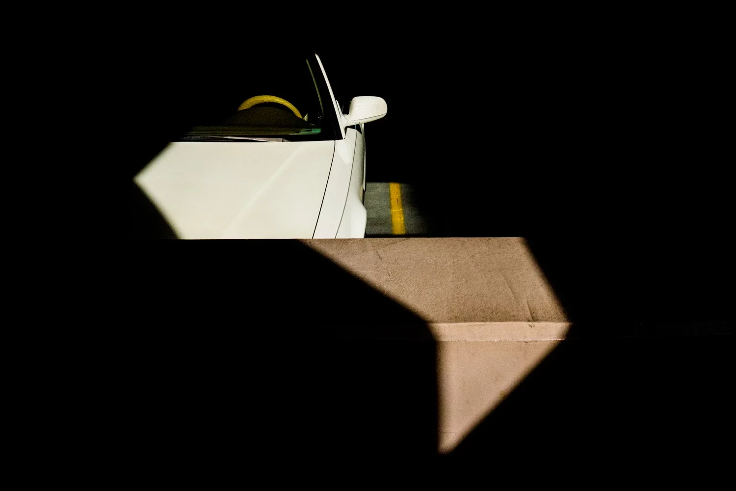 Part of a white car with a yellow item on the dashboard, seen through a small opening in a dark wall or structure, with a yellow line on the ground in the background.