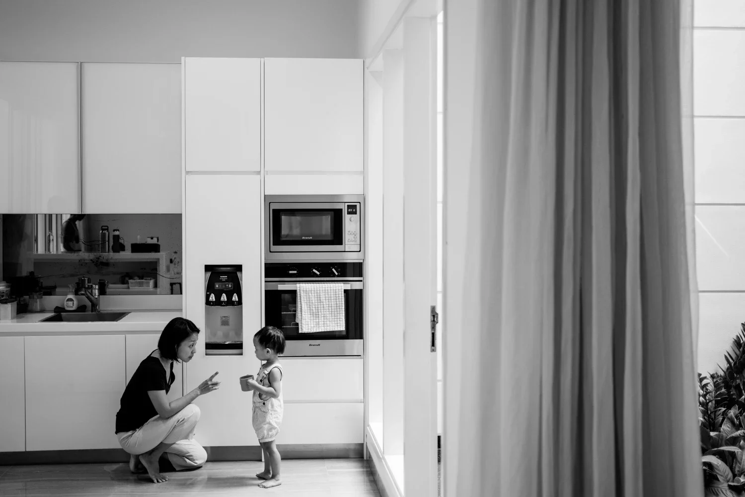 A woman kneeling and talking to a small child in a kitchen, with white cabinetry and built-in appliances, while holding a smartphone.
