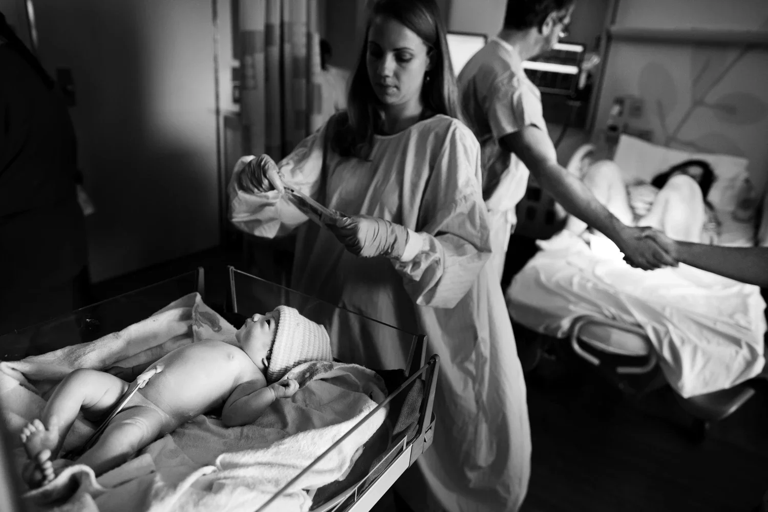 Newborn baby lying in a hospital bassinet with a hat, surrounded by medical staff and visitors in a hospital room.