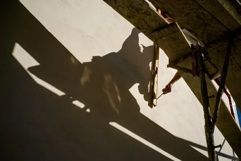 A silhouette of Kenton Nelson wearing a hat on top of scaffolding with a ruler and pencil, marking on a blank wall photographed by Erwin Darmali